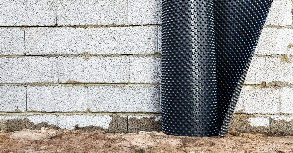 Close up view of a concrete block foundation wall with a roll of black dimpled drainage membrane partially unrolled beside it, for basement waterproofing in Hyattsville