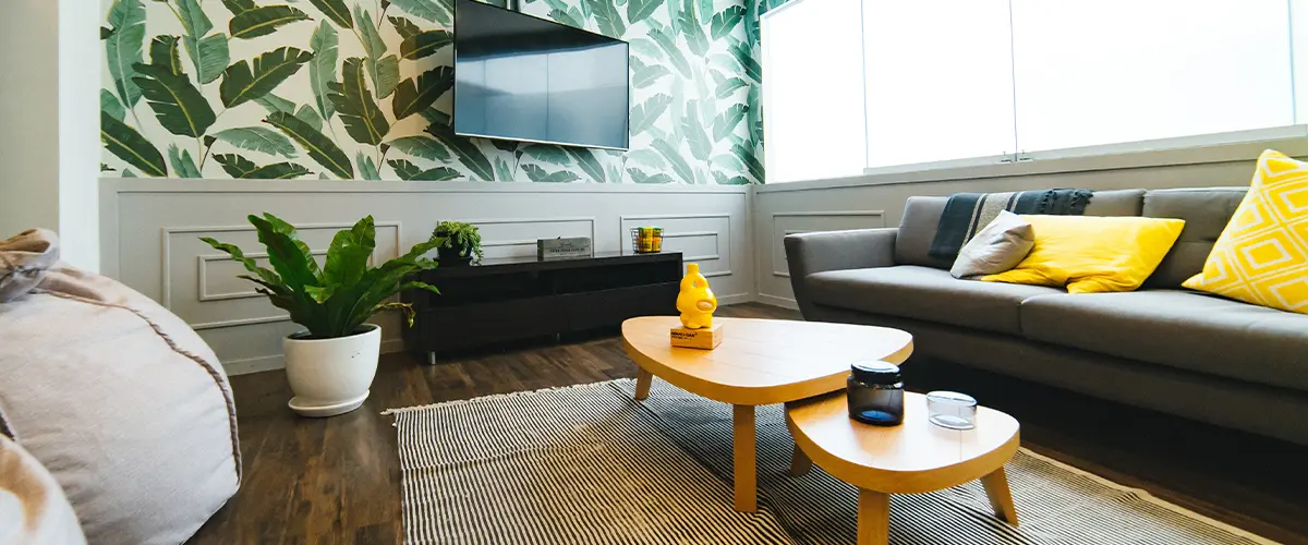 Modern living room with tropical leaf wallpaper, gray sofa with yellow pillows, wooden coffee table, and indoor plants.